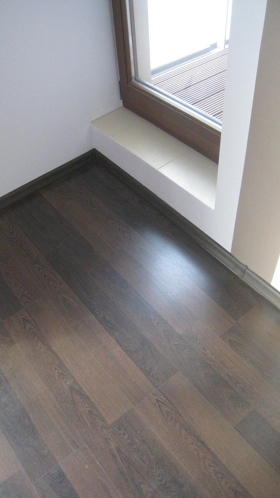 Ułożone panele podłogowe w odcieniach brązu w mieszkaniu w Warszawie, widoczne listwy przypodłogowe oraz fragment okna z parapetem.